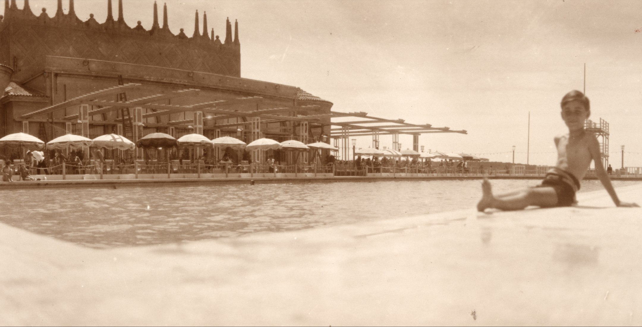 Early 20th century black and white photo of a young boy siting by a resort pool.