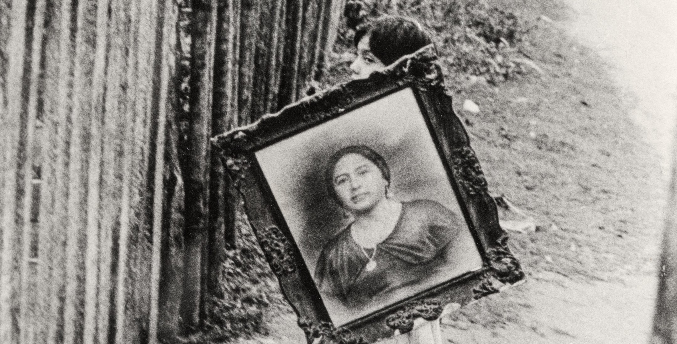 Black and white photo of a young girl carrying a framed portrait of a woman.