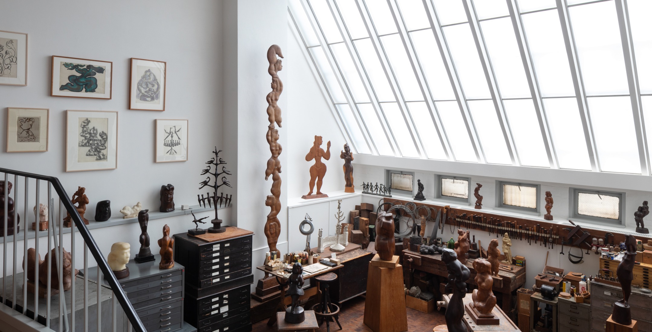 Photo of the studio at 526 LaGuardia Place. There is a skylight along the right half of the space that floods the sculptures, tools, and materials with light. There are drawings on the wall and many sculptures of human figures in wood.