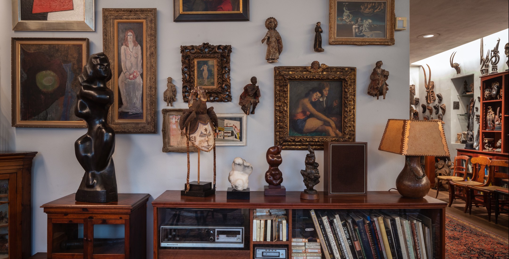 On a wooden table are various small sculptures and a brown lamp. On the grey wall are small mounted sculptures and various paintings of different sizes. The photo reveals a row of chairs and more mounted sculptures in the distance, around the corner.