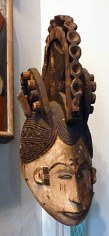 Polychrome wooden mask replicating the human profile. The features of the face are outlined in brown, including a six point star in between the figure's eyebrows. The hair is depicted in an elaborate composition of braids, kept close on the sides, while three large semicircle crests of wooden hair tower above the mask, like rooster combs.