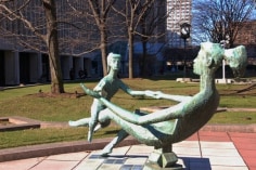 Green bronze sculpture in a park. The sculpture depicts a mother and child facing each other and playing.