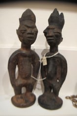 Two small twin sculptures with similar features placed next to each other. Both are made of dark wood and have their hair spiked up in a triangle fashion. The one on the left has a slightly smaller form, and both of them have the same closed eyes, wide, small nose, and circular mouths.