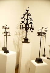 Six metal sculptures that resemble trees are mounted on rectangular, white boxes directly in front of us. At the middle is the largest sculpture, a tree with tiered branches with birds sitting upon them.