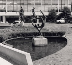 Black and white photograph of a sculpture in the middle of a small pond. The sculpture depicts a figure holding itself up on one hand, while balancing smaller figures on its foot in mid-air.