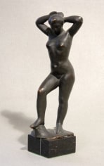 Bronze sculpture of a nude woman. She is standing on a stone platform, her right (viewer's left) leg slightly bent as it rests on a raised bronze surface emerging from the platform. Her head faces the floor and her hands rest on her head.