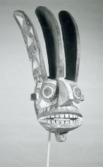Black and white photo of a wood and polychrome mask replicating a human profile, featuring exaggerated facial features, including widened and enlarged mouth and teeth, large projecting nose, and large eyes. Three curved vertical horns, carved in relief with geometric designs, emerge from the top of the mask.