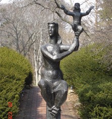 Bronze sculpture of a seated female figure. She is holding a smaller figure in her hands. The sculpture is placed in an outdoor setting by a brick path.