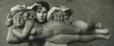 Black and white photo of Chaim Gross with a sculpture. The wood sculpture depicts a nude woman figure laying on her side with a small child's head and body resting behind her on her hips and legs. Chaim Gross is standing behind the sculpture, resting his arms on it and looking straight ahead.