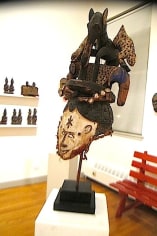 Wood mask mounted on a white block. The bottom part of the mask features a humanoid face in white, with brown outlining the facial features and black swirls representing the hair. On top of the face is a black, animal like creature with white and brown fabric surrounding it.