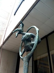 Close-up image of a bronze sculpture, turned green, in front of a building. The sculpture depicts a large figure and a small figure performing acrobatic tricks inside a circular hoop. The sculpture has a rough texture.