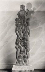 A black and white photo of a wood sculpture. The tall sculpture depicts multiple nude figures placed upon each other's shoulders.
