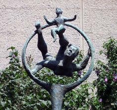 Dark green bronze sculpture of a figure leaping through a circular hoop. A smaller figure sits balanced on the larger figure's foot. The sculpture is rough in texture and is placed around flowers in an outdoor setting.