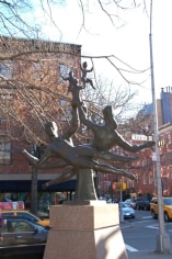 Color photograph of a sculpture in a park, with the city street in the background. The sculpture depicts several dancing figures leaping sideways through the air.