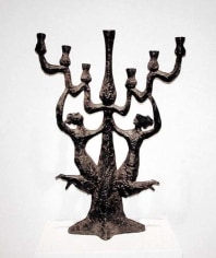 Dark-colored bronze menorah with a rough surface. Two female figures are attached to the stem of the menorah and are supporting the candle holders above.