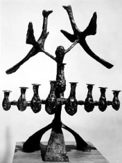 Image of a dark, sculpted menorah with the candle-holders placed in one row. Two sculpted birds seem to fly above the menorah.