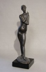 Bronze sculpture of a tall nude figure, made of metal. Its arms wrap around its chest and neck. It sits on a short stone base.