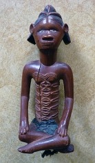 Wood, fabric, and raffia sculpture of a man wearing a cloth loincloth sitting with legs crossed. His elongated torso exhibits scarification.