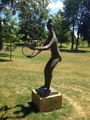 Image of a bronze sculpture in a sunny park. The sculpture is of a man juggling several circular hoops in his hands.