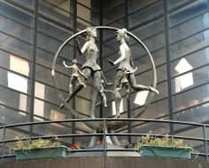 Sculpture of five figures dancing within a circular hoop. Each figure is facing a different direction. The sculpture is located in front of a building.