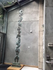 Image of a long, thin, vertical bronze sculpture. Each figure is stacked on top of the other, holding various poses.