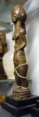 Wooden sculpture of the elongated and thin body of a nude woman, standing and holding her belly. Her body, especially her stomach, bears marks of scarification and she wears a long braid. Strands of white shells are tied around her neck, thighs, and ankles.