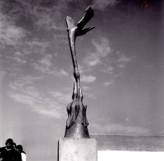 Black and white image of a sculpture in an outdoor setting. The sculpture depicts an abstract bird downward in flight, to feed smaller abstract birds reaching up at the base of the sculpture.