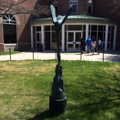 Image of a tall, thin sculpture on a patch of grass. The sculpture is of an abstract bird with its wings outstretched.