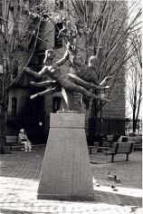 Black and white photograph of a sculpture in a park. The sculpture depicts several dancing figures leaping sideways through the air.