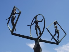 Looking up at this outdoor bronze sculpture, which has turned green, three figures stand with outstretched arms. The figures seem to be balancing on one leg, each one in the middle of their own circular or diamond shaped frame.
