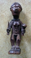 Wooden sculpture of a standing female figure, adorned with jewelry, and holding a knife (viewer's left) and staff or club (viewer's right) in each hand. The figure is presented with scarification marks on its torso, ivory inlaid eyes, and a metal ring around its neck.