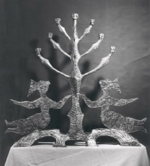 Black and white image of a bronze menorah. On each side of the menorah is a female figure with one arm reaching for the stem of the menorah and the other outstretched, while legs are outstretched on the base.