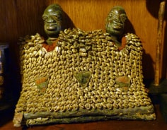 Wooden sculpture of twin figures, known as Ibeji, encased in square-like shawl of layered cowrie shells and beads. Only the heads of the figures are visible, emerging from a border of red fabric, framing their chests.
