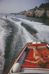 Slim Aarons, Leisure in Antibes, 1969: A woman sunbathes on a motorboat as it tows a waterskier in the bay off the Eden-Roc