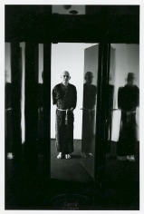 A black and white portrait of Michel Foucault in a robe, standing in a doorway. There is light in the hallway, his hands are behind his back.