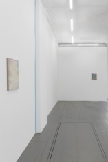 A photograph of the gallery's back quadrant where we see a pale painting by Monick in the foreground at left on the wall. Moving backward on that wall we see 2 ribbon works by Kovachevich (blue, green), then another Monick painting in the background at right.