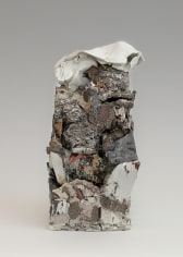 A sculpture of stacked stoneware. Several areas have a shiny silver luster. At the top is a folded piece of white ceramic that resembles a melted plastic bottle. There is a mixture of textures (gravel, sand, smooth stone) that resemble the environment despite being made entirely from clay.