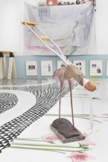 A photograph of a sculpture installed on the ground, made of a crutch attached to the body of a flamingo. In the background are a series of framed works hung low on the wall, contents illegible. There is also an artwork make of fabric above those framed works.