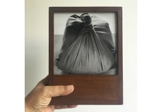 A hand holds a teak wood frame with a black and white photograph of a pothi, a knotted package made of fabric.