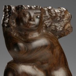 Rosewood sculpture of a crouched, sitting woman. Her head is cradled into her right bent elbow which rests on her knee while her other leg is tucked behind. Her hair flies back from her head, featuring pock mark textured detailing. 