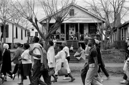 Selma to Montgomery civil rights marchers passing by house with people, African-American and white man holding small American flag in Montgomery, Alabama on March 25, 1965
