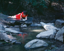 Rivers, First Draft: The Woman in Red, on a bed jutting into the water, skims an accordion-folded album of photos, 1982/2015, Digital C-print in 48 parts,&nbsp;16h x 20w in (40.64h x 50.80w cm)