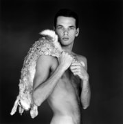 A nude young man from the waist up angling himself to look at the camera with a dead rabbit draped over his shoulder.
