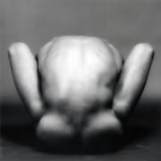 A nude figure crouching between his legs shown from behind.