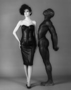 A woman in a leather cocktail dress and high heels facing the camera. An almost nude man stands next to her facing her and looking upward.