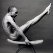 Naked man on stool with legs extended in a diagonal line.