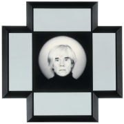 A portrait of the artist Andy Warhol from the neck up in a black turtleneck looking at the viewer. The black frame with white mat around the image forms a cross shape.