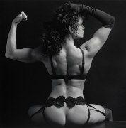 A woman in lingerie photographed from behind wearing one opera glove and flexing one arm.