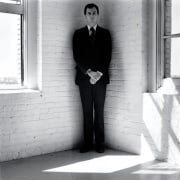 Robert Wilson wearing a suit standing in the corner of a white room.