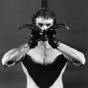 Shirtless man wearing leather gloves holding two horns in front of his face.